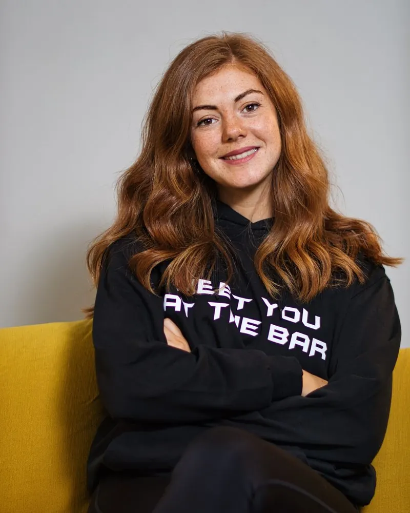 Mitarbeiterportrait einer lächelnden Frau mit verschränkten Armen auf einem gelben Sofa